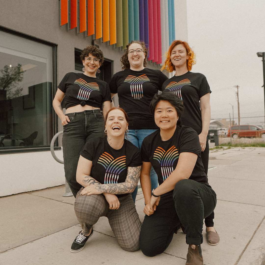 A group of OUTSaskatoon staff are standing and kneeling in front of their colourful office, smiling and all wearing this year's Hardpressed x OUTSaskatoon Pride rainbow sheaf t-shirt.