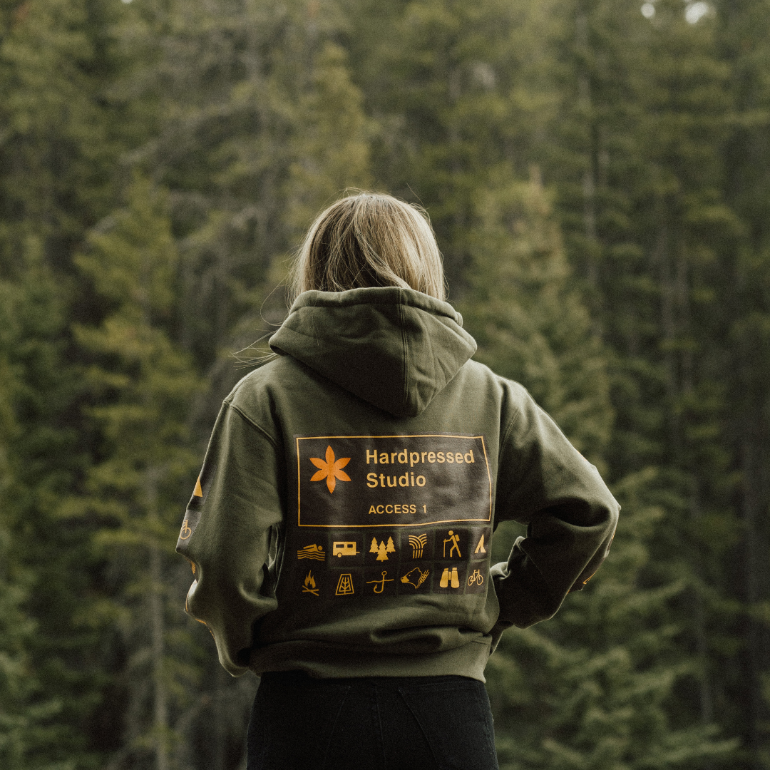 A woman wearing the Hardpressed Camp Series Sweater in Cypress green, facing the forest, showing the the printed camp sign graphic on the back of the hoodie.