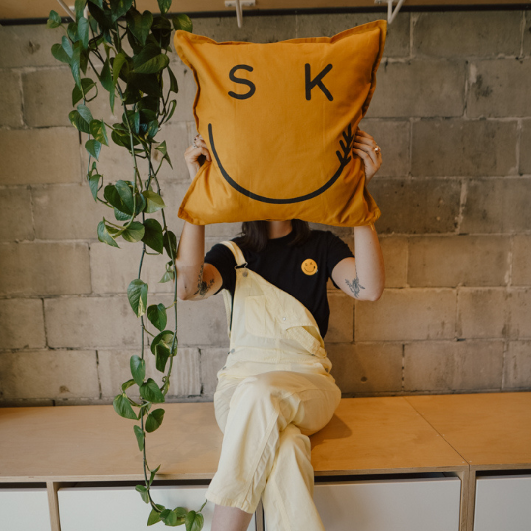 A woman is sitting on a bench with her legs crossed. She's wearing a black tee with a yellow crest smiley face graphic and yellow overalls. She's holding a smiley face pillow in front of her face.