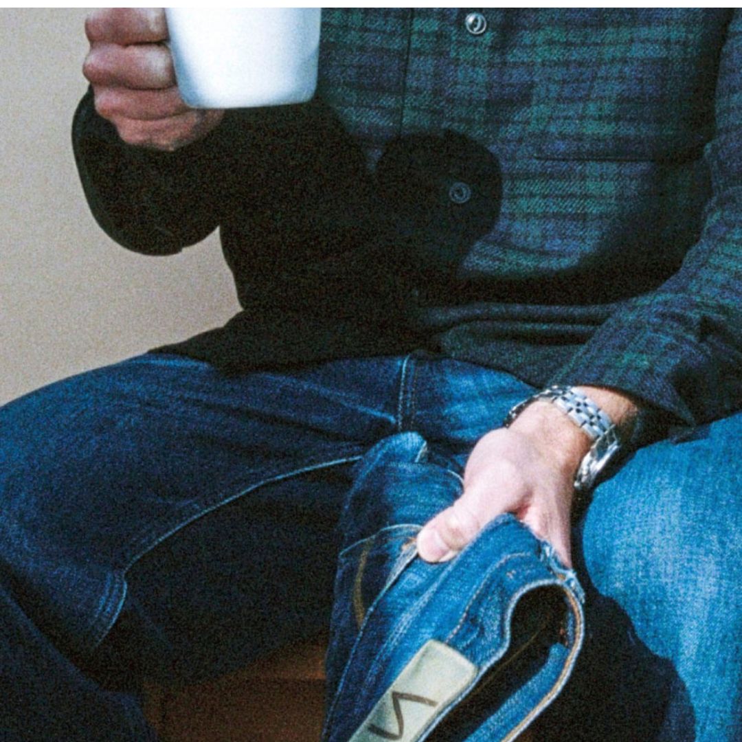 A man sitting down, holding a cup of coffee in one hand and a pair of Nudie Jeans in the other