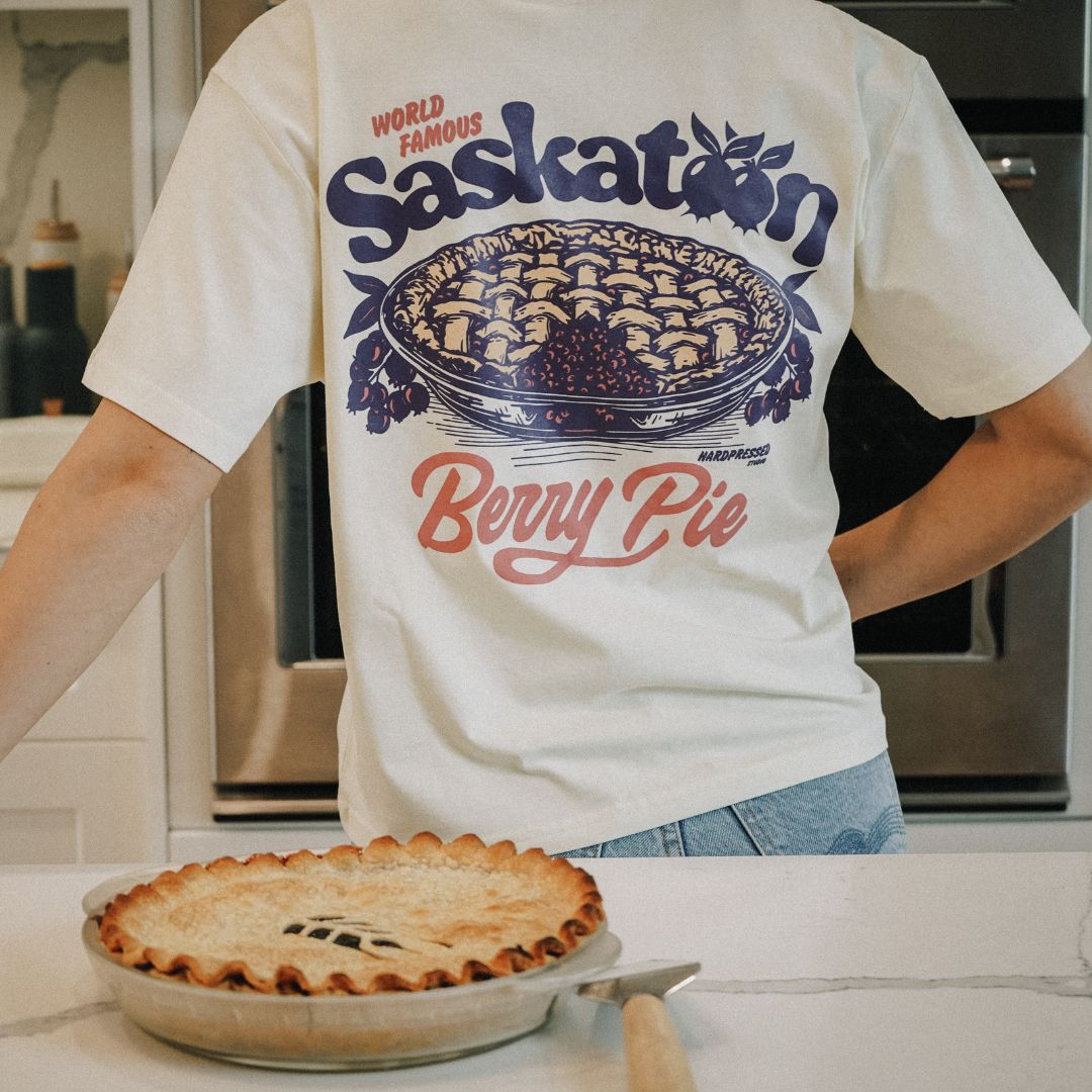 A pie sits on the counter in the foreground of a photo of Hardpressed's Saskatoon Berry Pie Tee. The photo shows the back of the graphic, which reads "World Famous Saskatoon Berry Pie" and has an image of a freshly baked pie with a slice out of it.